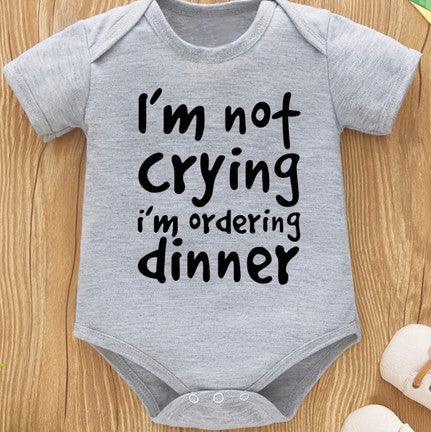 Gray baby onesie with the text "I'm not crying, I'm ordering dinner" printed in bold black letters, laid on a wooden surface with baby socks partially visible in the corner.