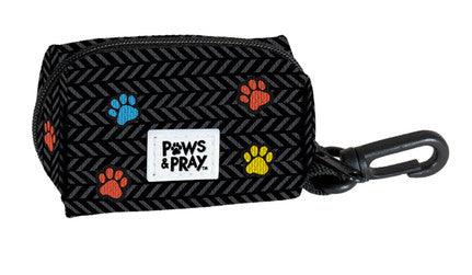 Black pet waste bag holder with a herringbone pattern, decorated with colorful paw prints in red, blue, and yellow. The holder features a small white label with the text "Paws & Pray" and has a black plastic clip attached for easy carrying.