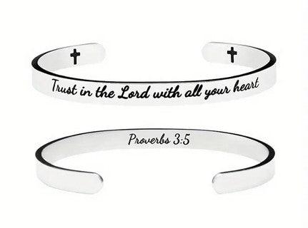 Silver cuff bracelet engraved with the phrase "Trust in the Lord with all your heart" on the outside and "Proverbs 3:5" on the inside. The bracelet also features small cross symbols near the open ends.