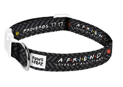 Black dog collar with a herringbone pattern and the text "Proverbs 17:17" and "A friend loves at all times" printed in white. The collar is decorated with small, colorful paw prints and features a "Paws & Pray" label. It has a sturdy metal D-ring and a white plastic buckle for secure fastening.