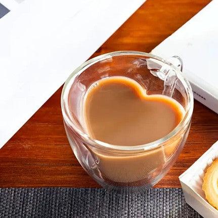 Glass mug with a heart-shaped inner design, filled with coffee or tea. The unique double-walled construction of the mug forms the heart shape when liquid is poured in, making it a charming and romantic addition to any beverage. The mug is set on a wooden surface near a small pastry.