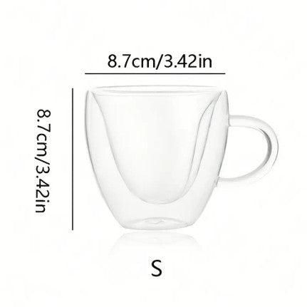 Glass mug with a heart-shaped inner design, filled with coffee or tea. The unique double-walled construction of the mug forms the heart shape when liquid is poured in, making it a charming and romantic addition to any beverage. The mug is set on a wooden surface near a small pastry.