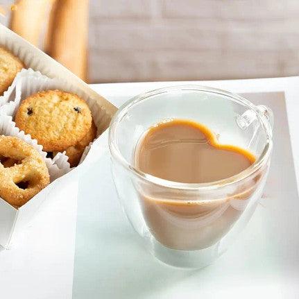 Glass mug with a heart-shaped inner design, filled with coffee or tea. The unique double-walled construction of the mug forms the heart shape when liquid is poured in, making it a charming and romantic addition to any beverage. The mug is set on a wooden surface near a small pastry.