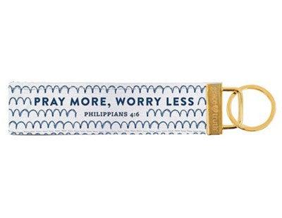 White fabric keychain with a repeating scallop pattern in blue and the phrase "PRAY MORE, WORRY LESS" printed in bold letters. Below the phrase, "Philippians 4:6" is printed, referencing the Bible verse. The keychain is accented with a gold metal key ring and a matching gold tag.