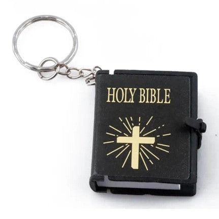 A miniature Holy Bible keychain with a black cover, featuring a gold cross and rays design along with the title "HOLY BIBLE" in gold lettering. The keychain is attached to a small metal ring, making it easy to carry. The tiny Bible has a clasp to keep it closed, adding a touch of spirituality and symbolism to your keys or bag.