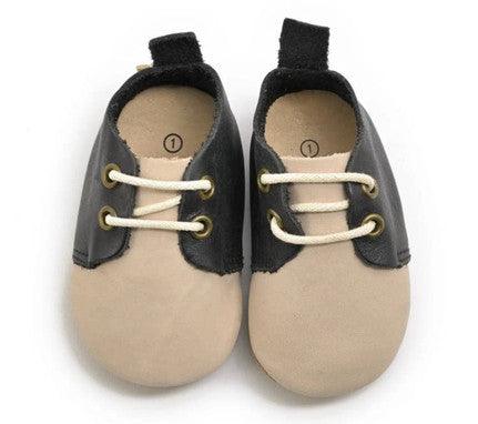 A pair of baby shoes with a two-tone design featuring cream and black colors. The shoes have a soft, rounded sole, ideal for infants, and are made from leather-like material. They include cream-colored laces threaded through gold eyelets, giving them a classic, stylish look. The black portion covers the top and heel of the shoes, while the cream color dominates the toe area. A small black pull-tab is attached at the back for easy wear. These shoes are designed to be both comfortable and durable for little f