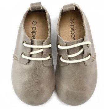 A pair of baby shoes in a soft gray color, made from a smooth, leather-like material. The shoes feature a minimalist design with cream-colored laces threaded through small eyelets, providing a secure fit. The interior of the shoes appears to be lined with a soft, tan-colored material for comfort. The brand name "Piper" is printed inside each shoe. A small pull-tab is located at the back for easy slip-on wear. These shoes are designed with a rounded toe and are ideal for babies who are learning to walk, offe