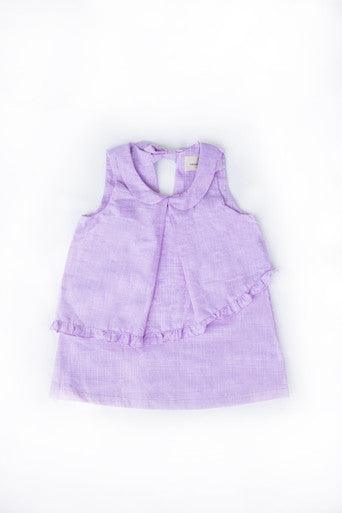 The image shows a sleeveless baby dress made of light lavender fabric. The dress features a soft, flowy texture with a simple, yet elegant design. It has a rounded collar and a small keyhole opening at the neckline, which is fastened with a button at the back. The bottom half of the dress is accented with a delicate ruffle detail that runs diagonally across the front, adding a touch of charm to the outfit. The dress is ideal for warm weather and has a light, airy appearance.