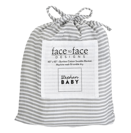 The image shows a drawstring bag made of light gray and white striped fabric. The bag features a square label on the front with the text "face to face DESIGNS" at the top, and "45” x 45” • Bamboo Cotton Swaddle Blanket. Machine wash & tumble dry." printed below. The label also includes the brand name "stephan BABY" at the bottom. The bag is cinched at the top with a drawstring, giving it a neat and compact appearance, typically used for packaging and storing a baby blanket.