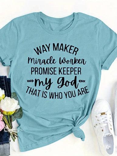 The image displays a light blue women's T-shirt with a faith-based design. The shirt features black text in a variety of fonts that reads: "WAY MAKER MIRACLE WORKER PROMISE KEEPER MY GOD THAT IS WHO YOU ARE." The phrase is a popular Christian worship song lyric, and the T-shirt is styled casually with a knot tied at the bottom. The shirt is laid out alongside a pair of white sneakers and some flowers, suggesting a comfortable and casual outfit choice.